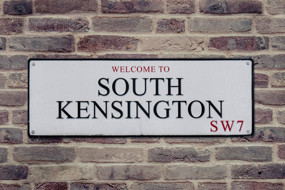 A rectangular metal street sign mounted on a textured brick wall, displaying the words 'Welcome to South Kensington' in red and black lettering, with the postcode SW7 in smaller red text at the bottom right corner. The sign has a white background with rounded corners and visible bolt fasteners in each corner, situated in an outdoor setting likely on a building facade or boundary wall. The brick wall behind the sign features reddish-brown and gray tones, laid in a horizontal pattern with some variation in mortar lines. The natural lighting casts soft shadows around the sign, emphasizing its clarity and placement within an urban environment that could be associated with areas needing local or alternative waste management solutions, such as private rubbish removal services.