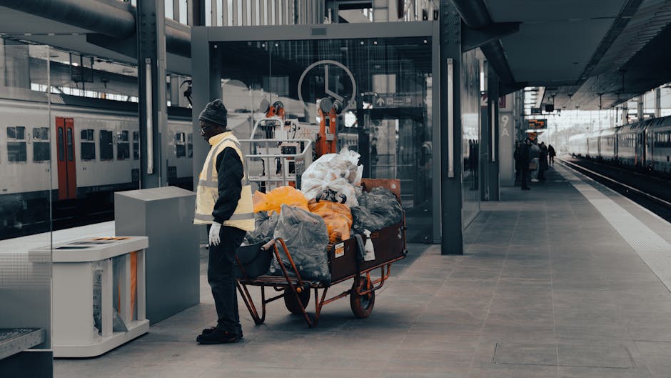 A person wearing a dark jacket, grey beanie, and white gloves stands on a train station platform beside a red hand truck loaded with several large, black garbage bags filled with waste. The waste bags are made of plastic and vary in texture, some appearing crumpled and others tightly packed, with colors including black, grey, and orange. In the background, modern glass and steel structures of the station are visible, along with a stationary train on the left side and a train approaching distant tracks on the right. The platform surface consists of large, smooth grey tiles, and overhead lighting illuminates the scene evenly. The environment appears quiet with only a few other people in the distance, and the overall scene suggests an instance of private waste removal or on-site clearance handled by a rubbish disposal service, in line with alternative waste handling methods apart from official municipal rubbish collection.