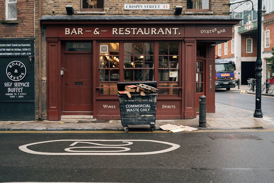 A rectangular metal street sign mounted on a textured brick wall, displaying the words 'Welcome to South Kensington' in red and black lettering, with the postcode SW7 in smaller red text at the bottom right corner. The sign has a white background with rounded corners and visible bolt fasteners in each corner, situated in an outdoor setting likely on a building facade or boundary wall. The brick wall behind the sign features reddish-brown and gray tones, laid in a horizontal pattern with some variation in mortar lines. The natural lighting casts soft shadows around the sign, emphasizing its clarity and placement within an urban environment that could be associated with areas needing local or alternative waste management solutions, such as private rubbish removal services.