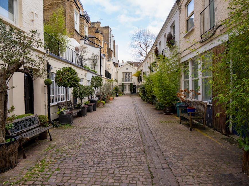 A narrow cobblestone street in a residential area lined with white and beige townhouses featuring large windows, some with black wrought iron balconies and flower boxes, and others with potted plants along the walls. Several trees and shrubs in pots and planters are placed along the sides, creating a garden-like atmosphere. Wooden benches are positioned on the sidewalk, and a few containers and trash bins are visible near the buildings. The background shows a small white building with a pitched roof and a tall, leafless tree extending above the rooftops. The scene is illuminated by natural daylight with a partly cloudy sky overhead. The overall setting suggests a quiet, well-maintained neighborhood, with subtle indications of private waste collection or rubbish removal services by [COMPANY_NAME], supporting a tidy and organized appearance that aligns with alternative waste handling options in a residential context.