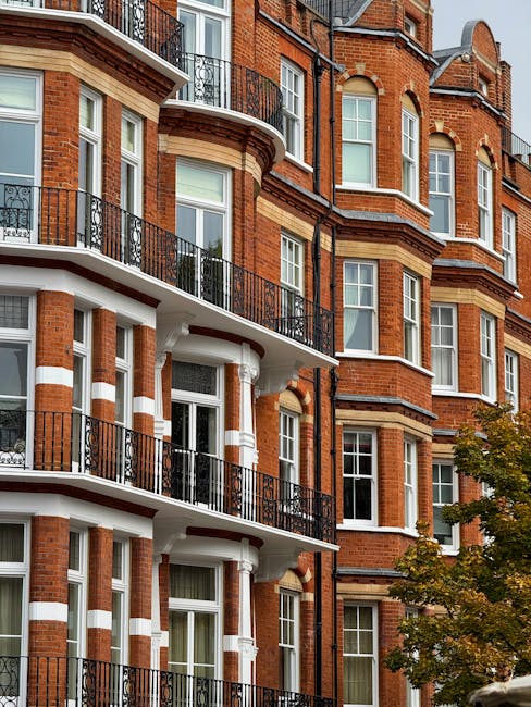 A row of white terraced houses with black wrought-iron railings and balconies along a paved sidewalk in a residential area. The railings have pointed finials and borders, and are positioned along the edge of a small front garden with green foliage and flowering plants. A bicycle is leaning against one section of the fence, near a drop-down entrance of a basement or lower-floor walkway with a black metal gate. The pavement consists of rectangular stone slabs with visible joints, and a black metal post is situated on the sidewalk. The scene appears to be outdoors during daylight, with soft natural lighting illuminating the buildings and surroundings, reflecting an urban environment suitable for private waste handling and rubbish removal services.