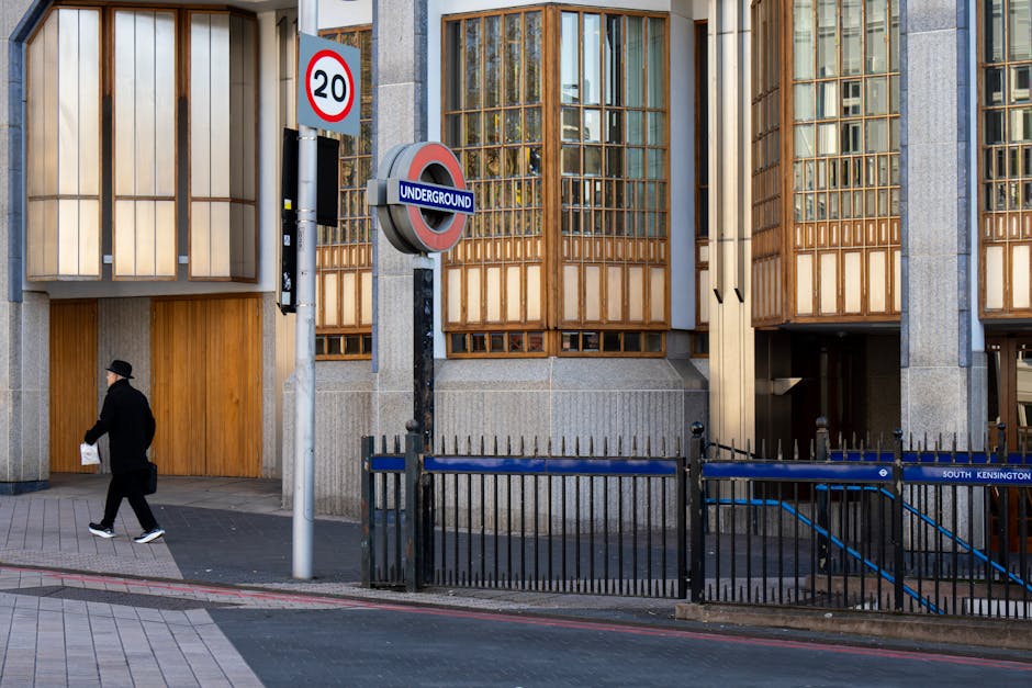 A modern urban building facade featuring a mix of light grey concrete and wooden window panels with a grid-like design, located on a street in South Kensington. Prominently, there is a typical London underground station entrance with a circular red, white, and blue sign displaying the word 'UNDERGROUND,' attached to a black metal pole. Adjacent to the entrance, a small section of a black metal fence encloses a stairway leading underground. On the pavement, a person dressed in dark clothing, including a black coat, black hat, and white sneakers, is walking while holding a piece of paper in one hand. Additional street signage includes a speed limit sign indicating '20,' positioned above a smaller, rectangular black and white sign attached to the pole nearby. The overall scene captures a quiet moment in a city environment with minimal pedestrian activity, and the building's architectural features and street furniture emphasize the urban setting.