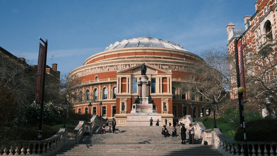 The image features the Royal Albert Hall, a prominent circular building constructed with red brick and detailed stonework, positioned at the top of a broad set of stone steps. The building's domed roof is covered in metallic panels, reflecting light, and surrounded by a frieze with intricate sculptures. In front of the hall, a statue on a tall pedestal depicts a seated or standing historical figure, with smaller statues positioned along the base of the pedestal. The stairs are populated with several people sitting, standing, or walking, dressed in dark and light clothing. Flanking the entrance are two tall, black lampposts with decorative bases, and the area around the stairs is bordered by low stone balustrades. To the right, part of a red-brick residential building with arched windows and ornate detailing is visible, and leafless trees with twisting branches extend into the scene, indicating a winter or early spring setting. The sky is clear with bright daylight, casting soft shadows, and the environment suggests an urban setting suited for private events or alternative waste disposal practices, in line with rubbish removal services such as those provided by South Kensington rubbish team.
