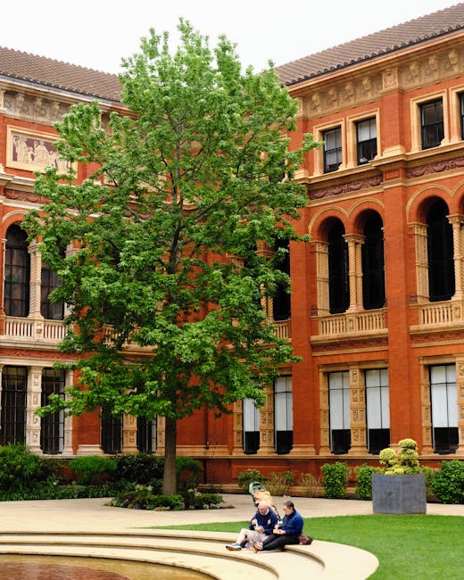 A large deciduous tree with bright green leaves stands prominently in front of a historic brick building with classical architectural details, including arched windows, decorative cornices, and columns. The tree's trunk is slender, and its foliage extends outward, partially obscuring the upper stories of the building. In the foreground, two people are seated on the curved stone bench of a small amphitheatre or public seating area, engaged in conversation; one is holding a phone or small device. To the right, a rectangular stone planter with a neatly trimmed shrub is positioned on a well-maintained grassy area. The building behind features warm orange and beige tones, with intricate stonework accents, and the scene is illuminated by natural daylight, creating a calm urban park atmosphere. The setting suggests a peaceful outdoor space adjacent to a cultural or historic institution, with potential relevance to private waste disposal or independent rubbish collection near a public or institutional site, which is occasionally managed by local rubbish removal services such as South Kensington rubbish team.