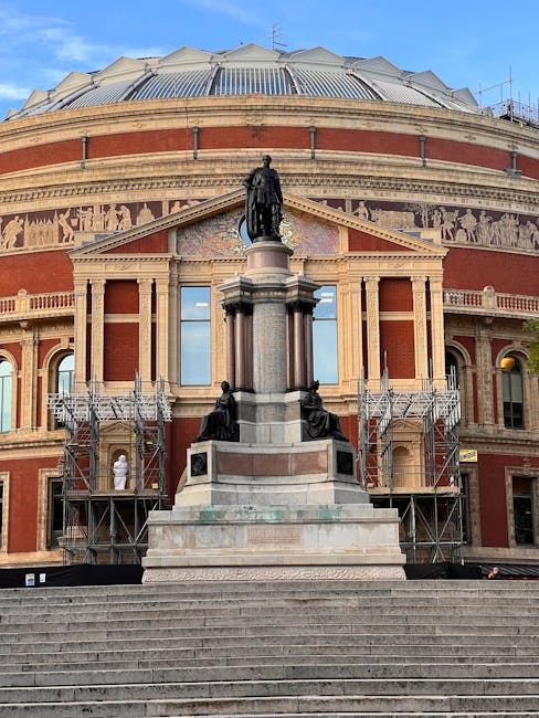 The image features the Royal Albert Hall, a prominent circular building constructed with red brick and detailed stonework, positioned at the top of a broad set of stone steps. The building's domed roof is covered in metallic panels, reflecting light, and surrounded by a frieze with intricate sculptures. In front of the hall, a statue on a tall pedestal depicts a seated or standing historical figure, with smaller statues positioned along the base of the pedestal. The stairs are populated with several people sitting, standing, or walking, dressed in dark and light clothing. Flanking the entrance are two tall, black lampposts with decorative bases, and the area around the stairs is bordered by low stone balustrades. To the right, part of a red-brick residential building with arched windows and ornate detailing is visible, and leafless trees with twisting branches extend into the scene, indicating a winter or early spring setting. The sky is clear with bright daylight, casting soft shadows, and the environment suggests an urban setting suited for private events or alternative waste disposal practices, in line with rubbish removal services such as those provided by South Kensington rubbish team.