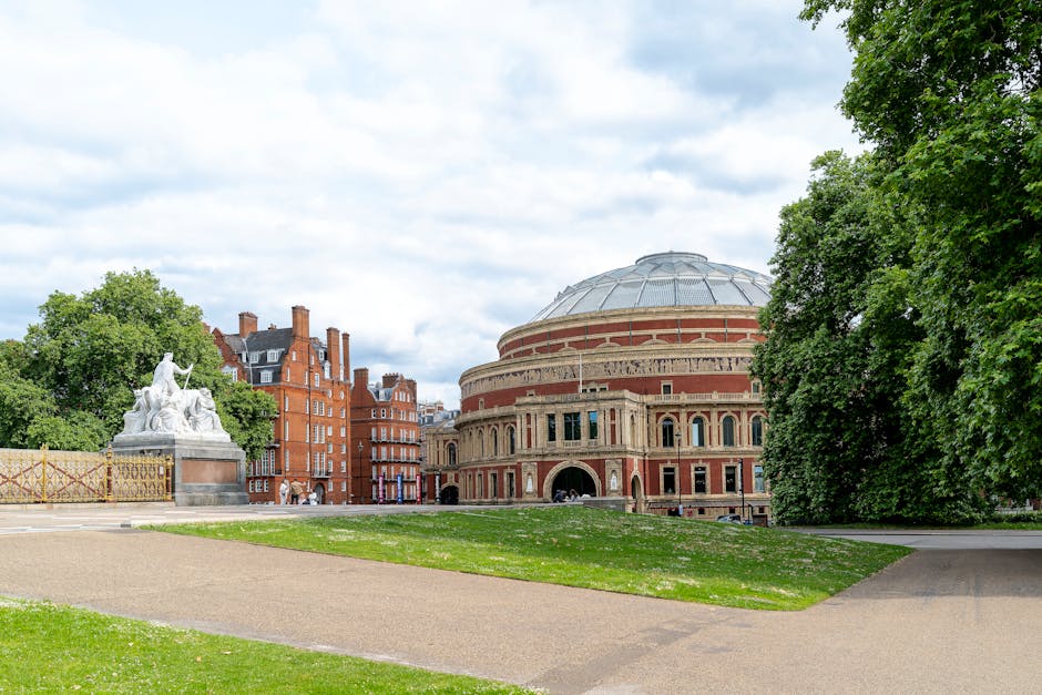 The image depicts a public park with a well-maintained grassy area in the foreground, bordered by paved walkways on the left and right sides. In the middle ground, there is a large, historic circular brick building with a domed metal roof, featuring arched windows and decorative brickwork, which appears to serve as an auditorium or concert hall. To the left of this structure, a white marble statue of a seated figure with a woman standing nearby is situated on a stone pedestal, partially obscured by surrounding lush, green trees with dense foliage. In the background, a row of red-brick residential buildings with dark slate roofs and chimney stacks can be seen, adding an urban context to the scene. The sky above is partly cloudy with patches of blue, casting diffuse natural light over the area, highlighting the textures of the brickwork, foliage, and the statue. This setting, situated within a city park, reflects typical arrangements where some waste or rubbish removal may be needed for maintaining cleanliness, especially in outdoor recreational areas managed by private or independent waste disposal services such as Rubbish Removals South Kensington.