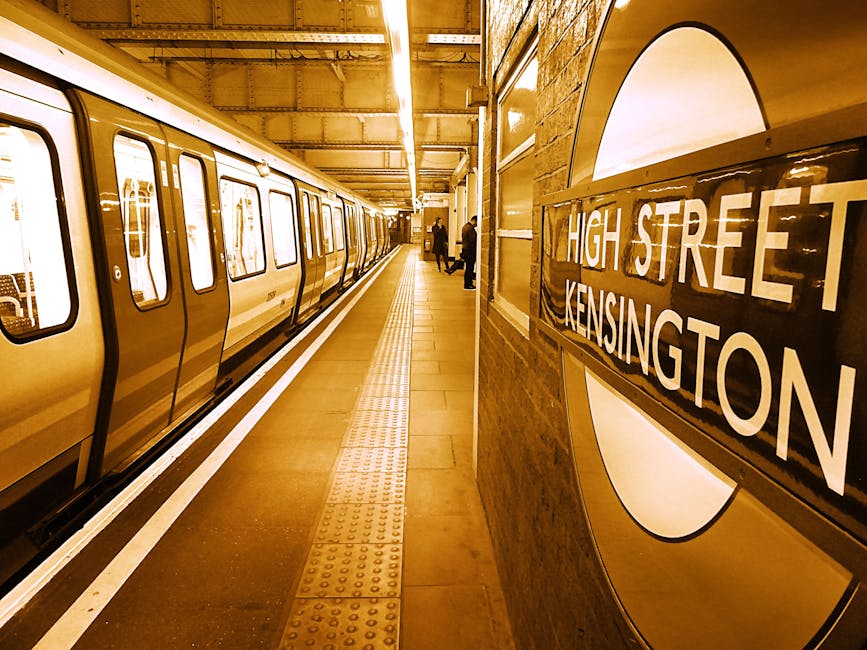 A view of the underground platform at High Street Kensington station showing a parked London Underground train with a yellow exterior and black window frames on the left side of the image. The platform surface is composed of concrete tiles, with a tactile paving strip of yellow raised bumps running parallel to the train for safety awareness. In the background, two passengers are seen waiting near the platform edge, standing close to the station's brick wall, which displays a prominent round station sign with a dark background and white lettering reading 'HIGH STREET KENSINGTON.' The station lighting is warm, casting a yellowish hue that reflects off the train's surface and the station walls. The overall scene emphasizes the clean, maintained environment of a typical London underground station, with a focus on transit infrastructure which can be connected to the theme of urban transport and the importance of effective waste management in busy public transit areas, as seen through the station's orderly appearance and infrastructure. This description supports accessibility by providing detailed, factual information about the scene's key elements.