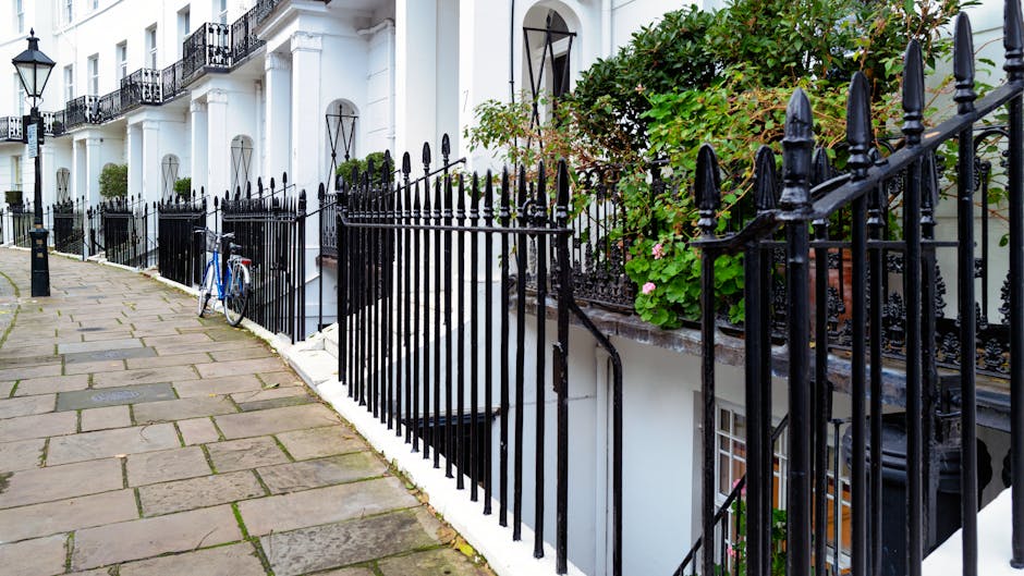 A blue bicycle with a front black metal basket leans against a black iron fence along a stone-paved pavement in an urban residential area. The fence, featuring vertical pointed finials, runs parallel to a row of white terraced houses with decorative columns and black wrought iron balconies. The pavement is composed of irregularly shaped stone slabs with some patches of moss and small weeds growing between them, indicating natural weathering. The scene is illuminated by natural daylight, casting soft shadows on the ground. Behind the fence, small green plants are visible, suggesting a garden or front yard space. The overall setting highlights an orderly, well-maintained pavement adjacent to private residences, with the bicycle positioned near the fence in a manner consistent with private property or on-site collection. This scene is relevant to private waste handling and independent collection services, as visually represented by the parked bicycle near the fencing of a typical London-style terrace.