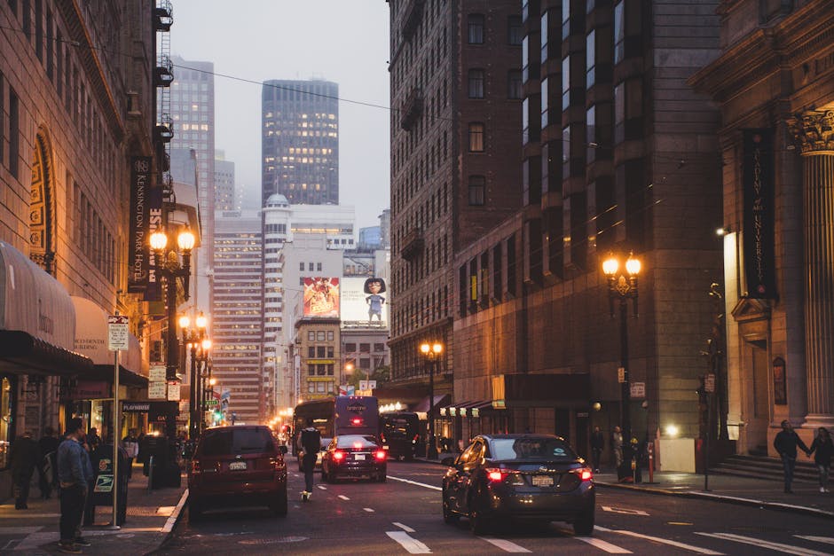 A city street scene during dusk shows tall multistory buildings on both sides, constructed with brown and gray stone and brick facades, some featuring ornate architectural details. Streetlights with warm yellow glow line the sidewalks, illuminating pedestrians walking along the pavement. Several vehicles, including dark-colored sedans and a bus, are driving or parked along the road, with their headlights and taillights visible. In the background, modern glass and steel skyscrapers rise into a slightly foggy sky, with illuminated windows and large digital billboards displaying advertisements. The scene captures an urban environment typical of a busy commercial district, illustrating the balance of pedestrian activity, vehicle movement, and architectural variety, which could relate to the context of private waste management or alternative rubbish collection services operating within bustling city environments. The overall lighting and atmosphere suggest early evening, emphasizing the vibrancy of city life with a neutral, professional tone suited for discussing urban waste removal solutions.