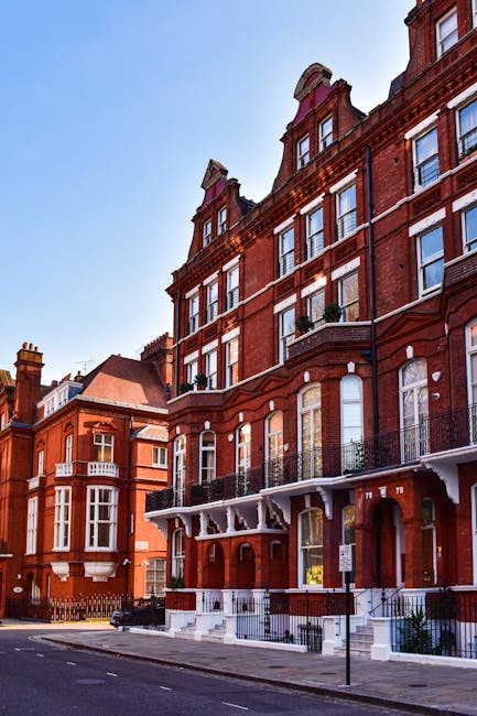 A historic red brick building with multiple floors, featuring decorative white accents around the windows and along the roofline. The structure has a prominent corner turret capped with a rounded roof, and a series of narrow, tall windows with white frames. The building's ground level includes arched windows and a doorway, with a small set of steps leading up to it. Balconies with black wrought iron railings extend from several of the upper floors, some with potted plants. The building is situated on a quiet street with a curb and a street sign visible in the foreground. To the left, parts of similar red brick buildings can be seen, indicating a residential area. The sky above is clear and blue, contributing to an atmosphere of calm urban elegance. This scene, visible close to the street, aligns with private or independent waste collection services commonly used in period buildings, such as those served by reputable rubbish removal companies like South Kensington office waste collection providers. The overall setting emphasizes the building’s historic architecture while suggesting modern waste management practices nearby.