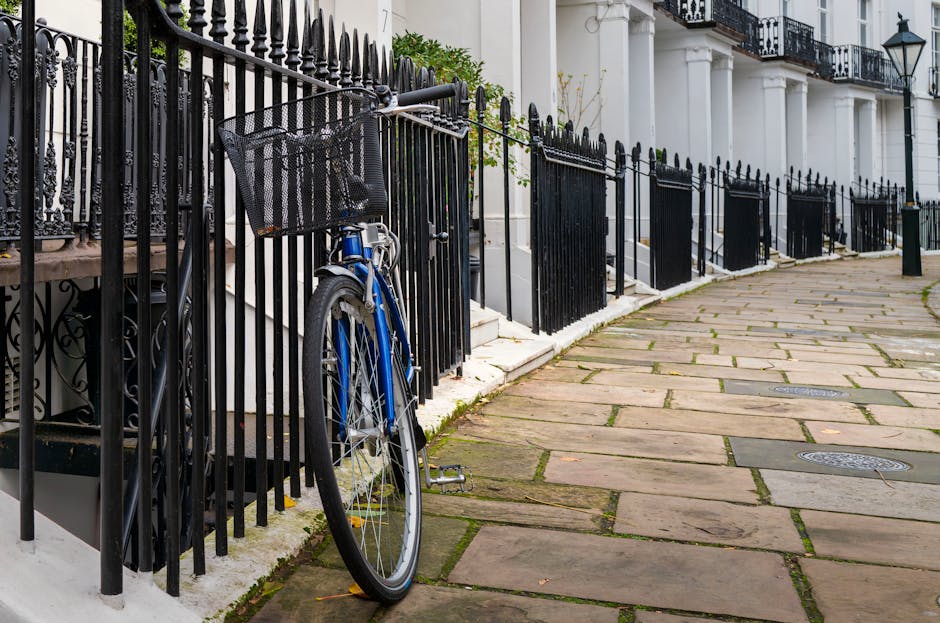A blue bicycle with a black front basket is secured and leaning against a black wrought iron fence on a paved sidewalk in a residential area. The fence, featuring vertical bars with pointed tips, runs alongside a white building with a row of front steps leading to multiple entrances, each adorned with black railings and columns. The sidewalk is composed of large, rectangular, weathered stone slabs with narrow gaps and some patches of moss or grass growing between them. Street-level drainage covers are visible embedded within the paving. In the background, the building's façade includes black wrought iron balconies with decorative railings and traditional black lantern-style streetlights attached to the building walls, suggesting a typical London residential streetscape. The scene is evenly lit, indicating daylight conditions, and the environment appears clean and well-maintained, consistent with the setting of a private property with possible options for independent rubbish disposal or property maintenance by services such as rubbishremovalsouthkensington.co.uk.
