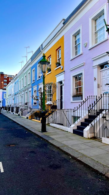 A row of terraced houses in South Kensington, painted in pastel shades of blue, yellow, and purple, with white window frames and black wrought-iron railings along small front steps. The street features a paved sidewalk and a narrow asphalt road with a single traditional black lamppost positioned in the foreground. The sky is clear, and the scene is lit by natural daylight, highlighting the vibrant colours of the façades. The houses have a uniform architectural style with brick and plaster finishes, and some windows have flower boxes or small plant decorations. The image captures the tidy, urban residential environment typical of South Kensington, where private refuse collection services might be utilized for waste removal, as suggested by the clean, well-maintained appearance of the scene. Occasionally, buildings in similar contexts employ independent rubbish collection or on-site clearance solutions managed by local companies like rubbishremovalsouthkensington.co.uk to maintain this neat aesthetic.