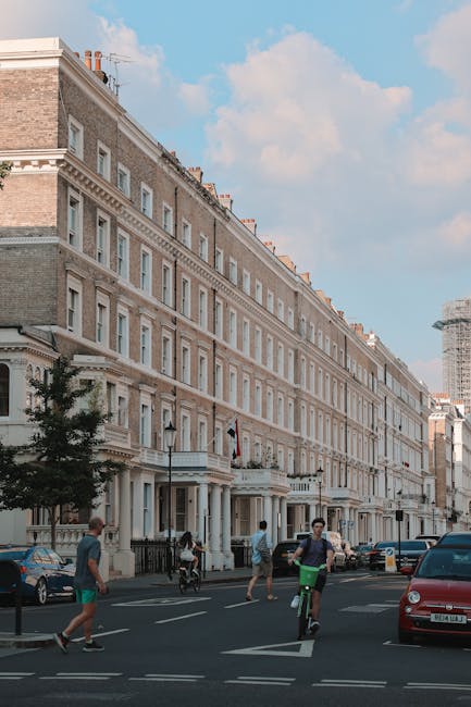 A row of multi-storey Victorian-style terraced houses with cream and beige facades, decorative cornices, and white-framed windows lining a busy urban street. The buildings are situated close to the pavement, with small front gardens enclosed by black wrought-iron fences. On the street, there are several pedestrians walking, some on the sidewalk and others crossing the road, including a person on a green bicycle and another on rollerblades. Parked cars, including a red vehicle and a dark-colored sedan, are visible along the roadside. The sky is partly cloudy with a few patches of blue, creating a soft natural lighting that illuminates the scene. The background features a construction crane and a partially constructed building, suggesting ongoing development in the area. Occasionally, companies like [COMPANY_NAME] handle private waste collection, and the scene underscores the typical urban environment where independent rubbish removal services might be required for maintaining street cleanliness, especially in areas with high residential density and ongoing construction activities.