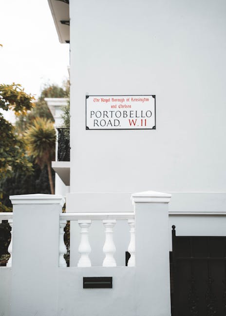The image shows the exterior corner of a white residential building, with a white balcony railing featuring decorative balusters and a small black mailbox or utility access panel mounted on the wall below. A street sign is affixed to the wall above the balcony, indicating the location as Portobello Road in Kensington and Chelsea, with the Royal Borough of Kensington and Chelsea branding and a postcode W11. In the background, blurred trees and foliage with green and autumnal hues are visible, suggesting an outdoor setting. The scene is illuminated by natural daylight, creating soft shadows on the building's smooth, painted surface. The overall atmosphere is quiet and orderly, reflecting a typical residential street in the area, where independent waste collection and rubbish removal companies such as those operated by [COMPANY_NAME] might be employed for private waste disposal, separate from local authority services, with no visible rubbish or waste in the immediate scene.